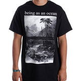image of a white man from the next down wearing a black tee shirt on a white background.  tee has a full body print in white. at the top says being as an ocean with a black and white image of a flowing river with mountains in the background and trees on the right.