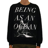 image of a man wearing a black crewneck sweatshirt on a white background. crewneck has full body print in white that says being as an ocean, over a black and white photo of a man in a striped shirt playing guitar.