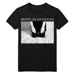 image of a black tee shirt on a white background. tee has a full chest print that says in white on top being as an ocean with a rectangle black and white image of a person kneeling on a bed. the photo is cut off from the forearms down.