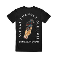 image of the back of a black tee shirt on a white background. the tee has a tan hand holding a blue and red bird tied in rope in the center with white text arhced around that says love has changed our lives and being as an ocean written below.
