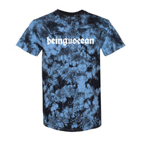 image of the front of a blue crystal wash tie dye tee shirt on a white background. the front of the tee has a white print across the chest that says being as an ocean
