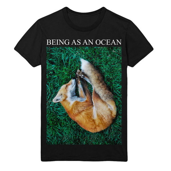 image of a black tee shirt on a white background. tee has full body print that says in white on top, being as an ocean with a photo of a sleeping fox on green grass. 