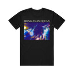 image of black tee shirt on a white background. tee has full chest print. in white on top says being as an ocean with a blurry image of a band playing with a crowd in blue and purple tones.