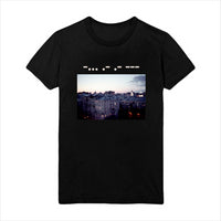 image of a black tee shirt on a white background. tee has white dashes and dots on the top in morse code with an image of rooftops at dusk below.