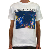 image of a white man wearing a white tee shirt on a white background. tee has full chest print. in black on top says being as an ocean with a blurry image of a band playing with a crowd in blue and purple tones.